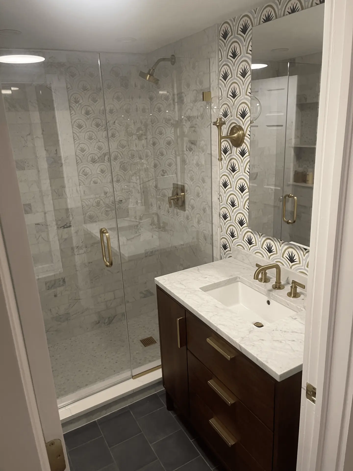 Renovated bathroom with a glass walk-in shower, patterned tile accent wall, walnut vanity, and brass fixtures